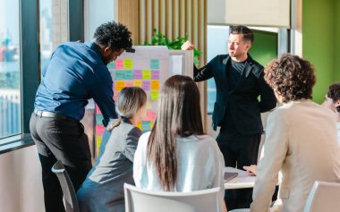 Çeşitli bir girişim ekibi, çevik bir görev tahtası kullanarak çalışma süreciyle ilgili bir tartışmaya girişir. Kurul, ekip planlama ve uygulama stratejilerinde işbirliği yaparken farklı proje aşamaları sergiliyor