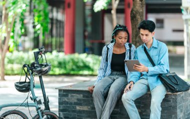 Üniversite öğrencileri iş projelerini tartışıyor. Bisiklet ve elektrikli scooter yakınlarda park edilmiş. Çevre dostu bir yol izlenimi uyandırıyor. Teknoloji ve sürdürülebilir ulaşım ile modern öğrenci yaşam tarzı