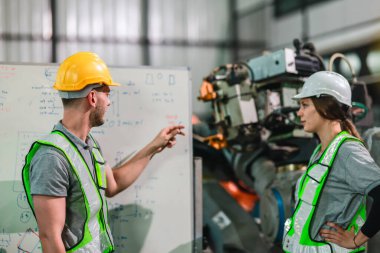 Güvenlik teçhizatı mühendisi ve teknisyeni modern bir fabrika ortamında robotik kol mekanizmalarını incelerken işbirliği yapıyorlar. Olay yeri takım çalışması, teknoloji ve ileri üretim süreçlerini vurguluyor