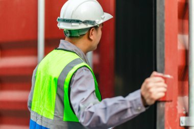Bir nakliye tesisinde konteynır teftişi yapan sanayi işçileri. Belgelerin incelenmesi ve lojistik, ulaşım ve kargo yönetiminde doğruluğu sağlamak için el bilgisayarı kullanılması.