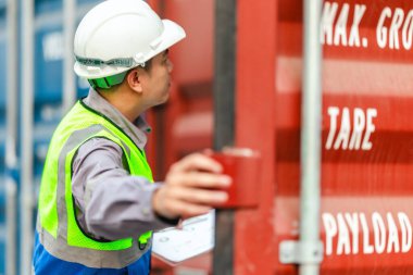Bir nakliye tesisinde konteynır teftişi yapan sanayi işçileri. Belgelerin incelenmesi ve lojistik, ulaşım ve kargo yönetiminde doğruluğu sağlamak için el bilgisayarı kullanılması.