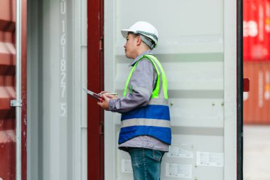 Bir nakliye tesisinde konteynır teftişi yapan sanayi işçileri. Belgelerin incelenmesi ve lojistik, ulaşım ve kargo yönetiminde doğruluğu sağlamak için el bilgisayarı kullanılması.