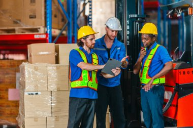 Güvenlik yelekleri ve miğferler içinde üç depo çalışanı, endüstriyel bir depolama tesisinde bir forklift ve bir kutu dolusu kutunun yanında tablet ve pano ile lojistiği koordine ediyorlar..