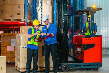 Güvenlik yelekleri ve miğferler içinde üç depo çalışanı, endüstriyel bir depolama tesisinde bir forklift ve bir kutu dolusu kutunun yanında tablet ve pano ile lojistiği koordine ediyorlar..