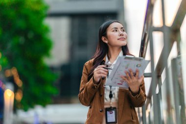 Kadın mimar, sürdürülebilir, çevre dostu ticari bina önündeki proje belgelerini inceliyor. Sahnede yeşil mimari, çevre sorumluluğu ve modern kentsel kalkınma vurgulanıyor