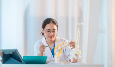 Asyalı tıp öğrencisi, ders kitabı ve dizüstü bilgisayarından bahsederken insan anatomik modellerini inceliyor, ayrıntılı bir çalışma oturumu, tıp eğitimi ve klinik ortamlarda elle öğrenme konularını vurguluyor.
