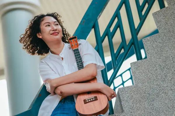 Kıvırcık saçlı Asyalı bir kadın açık hava ortamında ukulele çalıyor, mutluluk ve müzik enerjisi saçıyor. Doğal ve mimari unsurlara sahip rahat ve sanatsal bir ortam.