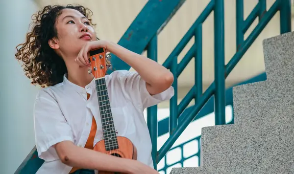 Kıvırcık saçlı Asyalı bir kadın açık hava ortamında ukulele çalıyor, mutluluk ve müzik enerjisi saçıyor. Doğal ve mimari unsurlara sahip rahat ve sanatsal bir ortam.