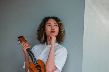 Kıvırcık saçlı Asyalı bir kadın açık hava ortamında ukulele çalıyor, mutluluk ve müzik enerjisi saçıyor. Doğal ve mimari unsurlara sahip rahat ve sanatsal bir ortam.