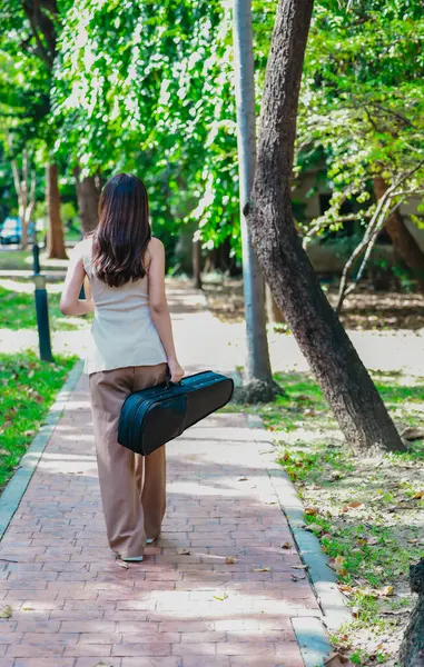 Uzun saçlı genç bir Asyalı kadın, elinde siyah bir keman çantasıyla güneşli bir parkta yürüyor..