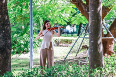 Genç bir Asyalı kadın bereketli yeşilliklerle çevrili huzurlu bir açık hava ortamında kibarca keman çalıyor. Doğada bir müzik konsantrasyonu ve sükunet anı yakalıyorum..