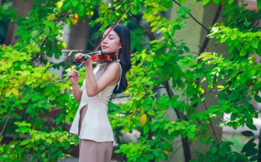 Genç bir Asyalı kadın bereketli yeşilliklerle çevrili huzurlu bir açık hava ortamında kibarca keman çalıyor. Doğada bir müzik konsantrasyonu ve sükunet anı yakalıyorum..
