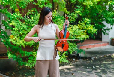 Genç bir Asyalı kadın bereketli yeşilliklerle çevrili huzurlu bir açık hava ortamında kibarca keman çalıyor. Doğada bir müzik konsantrasyonu ve sükunet anı yakalıyorum..