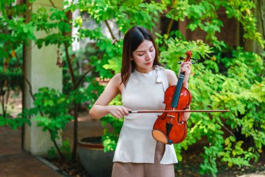 Genç bir Asyalı kadın bereketli yeşilliklerle çevrili huzurlu bir açık hava ortamında kibarca keman çalıyor. Doğada bir müzik konsantrasyonu ve sükunet anı yakalıyorum..