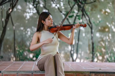 Genç bir Asyalı kadın bereketli yeşilliklerle çevrili huzurlu bir açık hava ortamında kibarca keman çalıyor. Doğada bir müzik konsantrasyonu ve sükunet anı yakalıyorum..