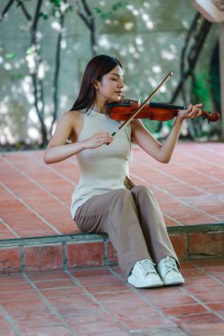 Genç bir Asyalı kadın bereketli yeşilliklerle çevrili huzurlu bir açık hava ortamında kibarca keman çalıyor. Doğada bir müzik konsantrasyonu ve sükunet anı yakalıyorum..