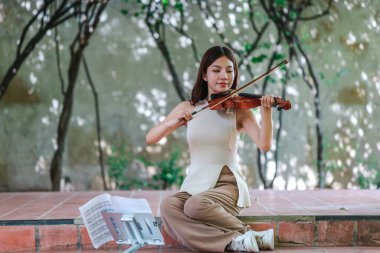 Genç bir Asyalı kadın bereketli yeşilliklerle çevrili huzurlu bir açık hava ortamında kibarca keman çalıyor. Doğada bir müzik konsantrasyonu ve sükunet anı yakalıyorum..
