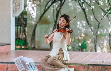 Genç bir Asyalı kadın bereketli yeşilliklerle çevrili huzurlu bir açık hava ortamında kibarca keman çalıyor. Doğada bir müzik konsantrasyonu ve sükunet anı yakalıyorum..