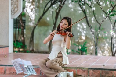 Genç bir Asyalı kadın bereketli yeşilliklerle çevrili huzurlu bir açık hava ortamında kibarca keman çalıyor. Doğada bir müzik konsantrasyonu ve sükunet anı yakalıyorum..