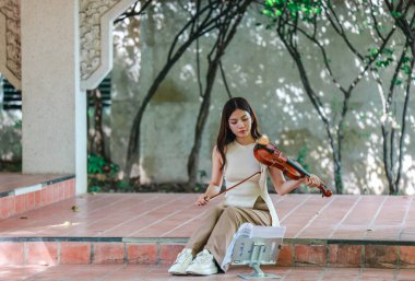 Genç bir Asyalı kadın bereketli yeşilliklerle çevrili huzurlu bir açık hava ortamında kibarca keman çalıyor. Doğada bir müzik konsantrasyonu ve sükunet anı yakalıyorum..