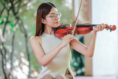Genç bir Asyalı kadın bereketli yeşilliklerle çevrili huzurlu bir açık hava ortamında kibarca keman çalıyor. Doğada bir müzik konsantrasyonu ve sükunet anı yakalıyorum..