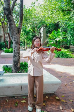 Genç bir Asyalı kadın bereketli yeşilliklerle çevrili huzurlu bir açık hava ortamında kibarca keman çalıyor. Doğada bir müzik konsantrasyonu ve sükunet anı yakalıyorum..