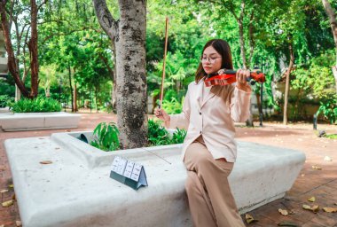 Genç bir Asyalı kadın bereketli yeşilliklerle çevrili huzurlu bir açık hava ortamında kibarca keman çalıyor. Doğada bir müzik konsantrasyonu ve sükunet anı yakalıyorum..