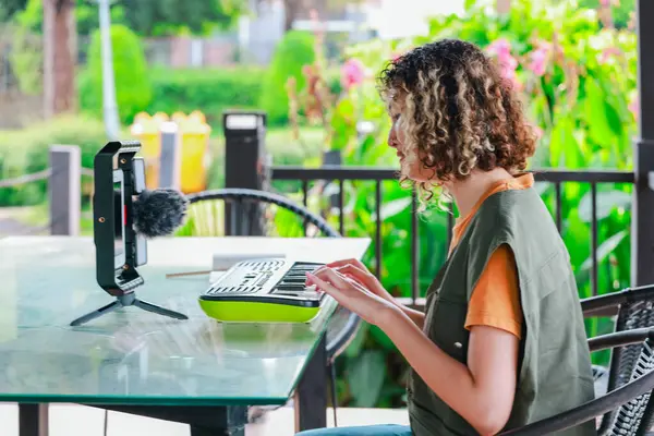 Kıvırcık saçlı Asyalı kadın çiçeklerle dolu bir bahçede küçük bir klavye ve akıllı telefon kullanarak çevrimiçi müzik seansına katılıyor. Açık havada yaratıcı bir öğrenme anı