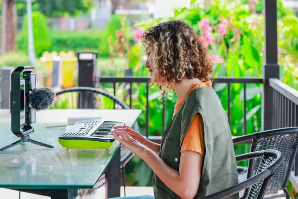 Kıvırcık saçlı Asyalı kadın çiçeklerle dolu bir bahçede küçük bir klavye ve akıllı telefon kullanarak çevrimiçi müzik seansına katılıyor. Açık havada yaratıcı bir öğrenme anı