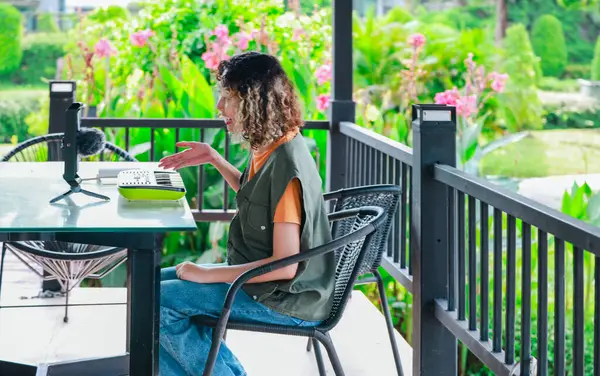 Kıvırcık saçlı Asyalı kadın çiçeklerle dolu bir bahçede küçük bir klavye ve akıllı telefon kullanarak çevrimiçi müzik seansına katılıyor. Açık havada yaratıcı bir öğrenme anı