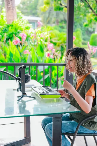 Kıvırcık saçlı Asyalı kadın çiçeklerle dolu bir bahçede küçük bir klavye ve akıllı telefon kullanarak çevrimiçi müzik seansına katılıyor. Açık havada yaratıcı bir öğrenme anı