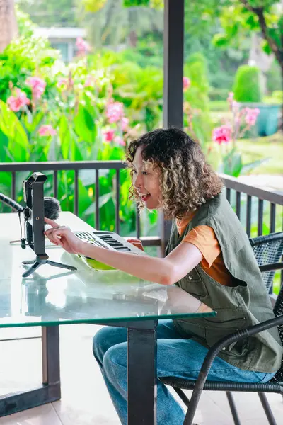 Kıvırcık saçlı Asyalı kadın çiçeklerle dolu bir bahçede küçük bir klavye ve akıllı telefon kullanarak çevrimiçi müzik seansına katılıyor. Açık havada yaratıcı bir öğrenme anı
