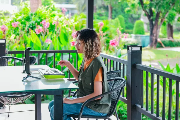 Kıvırcık saçlı Asyalı kadın çiçeklerle dolu bir bahçede küçük bir klavye ve akıllı telefon kullanarak çevrimiçi müzik seansına katılıyor. Açık havada yaratıcı bir öğrenme anı