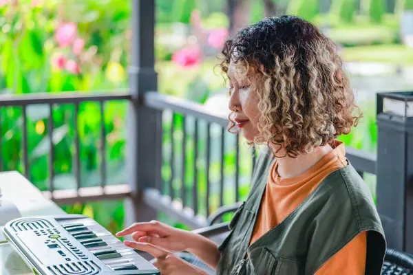 Kıvırcık saçlı Asyalı kadın çiçeklerle dolu bir bahçede küçük bir klavye ve akıllı telefon kullanarak çevrimiçi müzik seansına katılıyor. Açık havada yaratıcı bir öğrenme anı