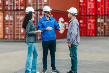 Kadın yönetici, konteyner deposunda iki erkek işçiyle lojistiği denetliyor ve tartışıyor. Küresel ticaret ve ulaşım endüstrisinin altını çizmek için toplanmış konteynırlar ve endüstriyel ekipmanlar