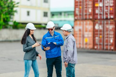 Kadın yönetici, konteyner deposunda iki erkek işçiyle lojistiği denetliyor ve tartışıyor. Küresel ticaret ve ulaşım endüstrisinin altını çizmek için toplanmış konteynırlar ve endüstriyel ekipmanlar