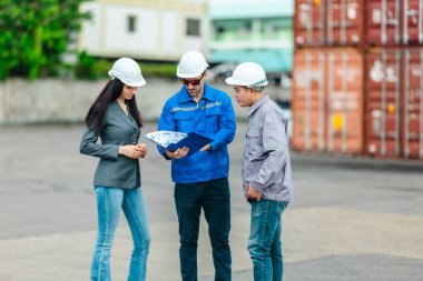 Kadın yönetici, konteyner deposunda iki erkek işçiyle lojistiği denetliyor ve tartışıyor. Küresel ticaret ve ulaşım endüstrisinin altını çizmek için toplanmış konteynırlar ve endüstriyel ekipmanlar