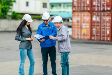Kadın yönetici, konteyner deposunda iki erkek işçiyle lojistiği denetliyor ve tartışıyor. Küresel ticaret ve ulaşım endüstrisinin altını çizmek için toplanmış konteynırlar ve endüstriyel ekipmanlar
