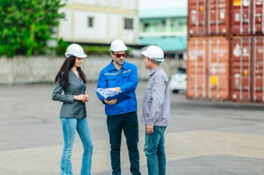 Kadın yönetici, konteyner deposunda iki erkek işçiyle lojistiği denetliyor ve tartışıyor. Küresel ticaret ve ulaşım endüstrisinin altını çizmek için toplanmış konteynırlar ve endüstriyel ekipmanlar