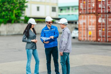 Kadın yönetici, konteyner deposunda iki erkek işçiyle lojistiği denetliyor ve tartışıyor. Küresel ticaret ve ulaşım endüstrisinin altını çizmek için toplanmış konteynırlar ve endüstriyel ekipmanlar