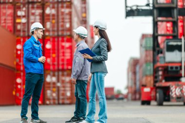 Kadın yönetici, konteyner deposunda iki erkek işçiyle lojistiği denetliyor ve tartışıyor. Küresel ticaret ve ulaşım endüstrisinin altını çizmek için toplanmış konteynırlar ve endüstriyel ekipmanlar