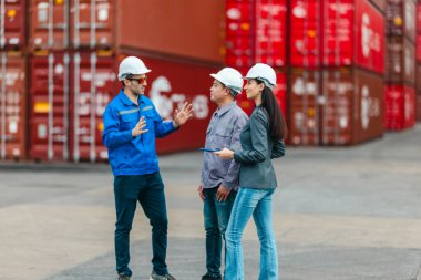 Kadın yönetici, konteyner deposunda iki erkek işçiyle lojistiği denetliyor ve tartışıyor. Küresel ticaret ve ulaşım endüstrisinin altını çizmek için toplanmış konteynırlar ve endüstriyel ekipmanlar