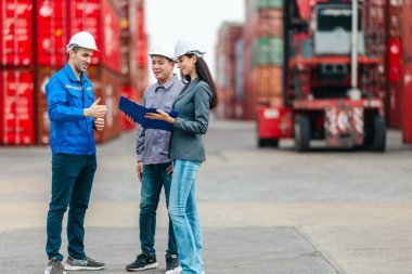 Kadın yönetici, konteyner deposunda iki erkek işçiyle lojistiği denetliyor ve tartışıyor. Küresel ticaret ve ulaşım endüstrisinin altını çizmek için toplanmış konteynırlar ve endüstriyel ekipmanlar