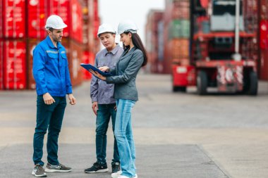 Kadın yönetici, konteyner deposunda iki erkek işçiyle lojistiği denetliyor ve tartışıyor. Küresel ticaret ve ulaşım endüstrisinin altını çizmek için toplanmış konteynırlar ve endüstriyel ekipmanlar