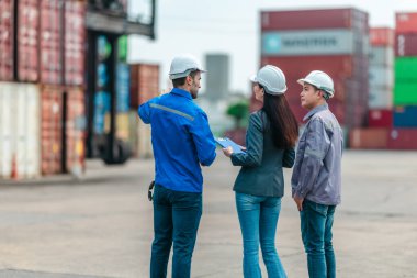 Kadın yönetici, konteyner deposunda iki erkek işçiyle lojistiği denetliyor ve tartışıyor. Küresel ticaret ve ulaşım endüstrisinin altını çizmek için toplanmış konteynırlar ve endüstriyel ekipmanlar