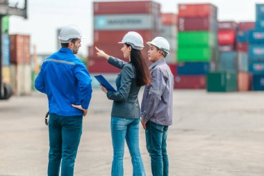 Kadın yönetici, konteyner deposunda iki erkek işçiyle lojistiği denetliyor ve tartışıyor. Küresel ticaret ve ulaşım endüstrisinin altını çizmek için toplanmış konteynırlar ve endüstriyel ekipmanlar