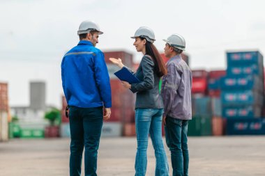 Kadın yönetici, konteyner deposunda iki erkek işçiyle lojistiği denetliyor ve tartışıyor. Küresel ticaret ve ulaşım endüstrisinin altını çizmek için toplanmış konteynırlar ve endüstriyel ekipmanlar