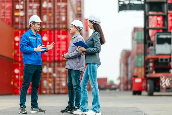 Kadın yönetici, konteyner deposunda iki erkek işçiyle lojistiği denetliyor ve tartışıyor. Küresel ticaret ve ulaşım endüstrisinin altını çizmek için toplanmış konteynırlar ve endüstriyel ekipmanlar