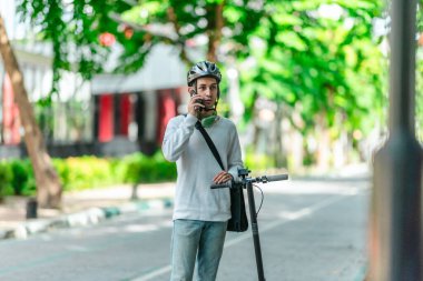 Kasklı genç bir adam, şehir caddesinde elektrikli scooterla dikilirken telefonuyla konuşuyor, modern, çevre dostu şehir ulaşımının özünü yakalıyor..