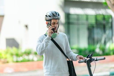 Kasklı genç bir adam, şehir caddesinde elektrikli scooterla dikilirken telefonuyla konuşuyor, modern, çevre dostu şehir ulaşımının özünü yakalıyor..